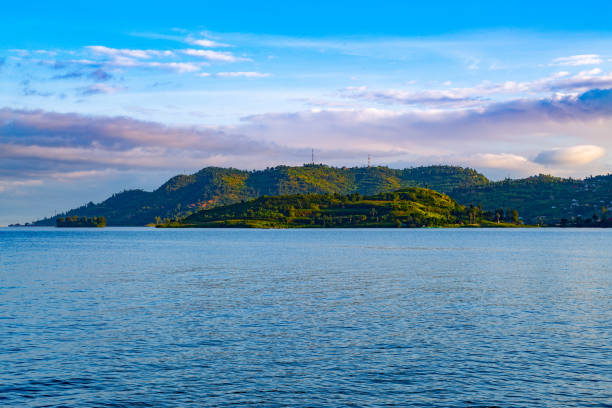 A tranquil view of Lake Kivu in Rwanda under a bright blue sky, with lush green hills in the background. The calm waters and soft clouds create a peaceful ambiance, capturing the beauty of this natural landscape at sunrise. Ideal for projects related to travel, nature, tranquility, and the scenic beauty of Rwanda.
